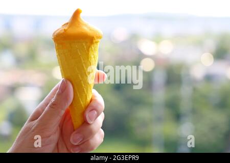 Eiscreme im Waffelkegel in weiblicher Hand im Hintergrund der Sommerstadt. Kühles Dessert mit Mangosaft bei heißem Wetter Stockfoto