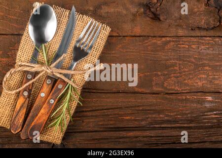 Besteck mit Leinenserviette und frischer Rosmarin-Dekoration auf rustikalem Holzsockel. Ein Löffel, eine Gabel und ein Messer als Restaurant- und Küchenutensilien. Stockfoto