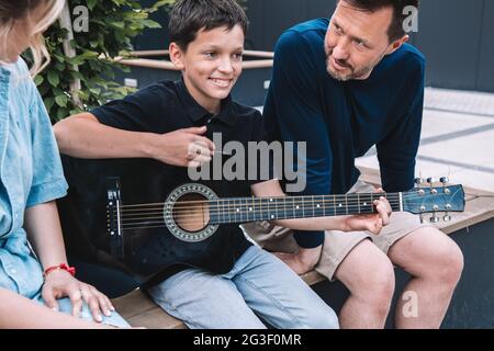 Eltern hören aufmerksam zu, während ihr Sohn die ersten Schritte beim Gitarrenspiel unternimmt. Sie sitzen am Sommertag auf der Bank auf dem Stadtplatz. Lifestyle. Hoch Stockfoto