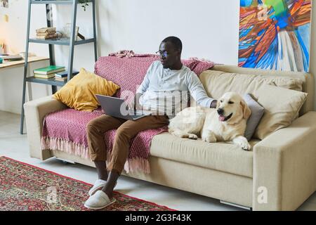 Afrikanischer Mann, der mit seinem Hund auf dem Sofa sitzt und seinen Laptop für die Online-Arbeit im heimischen Zimmer benutzt Stockfoto