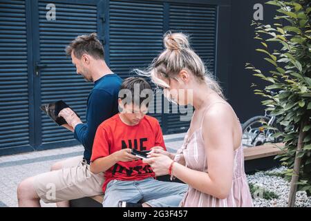 Die junge Familie mit ihrem Sohn sitzt auf der Bank auf dem Stadtplatz, alle mit digitalen Technologien Handys und Tablets . Sie kommunizieren nicht mit wi Stockfoto