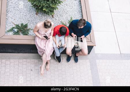 Vater, Mutter und Sohn in gewöhnlichen Kleidern sitzen auf der Bank auf der Straße, jeder nutzt Smartphone oder Tablet. Lifestyle. Stockfoto