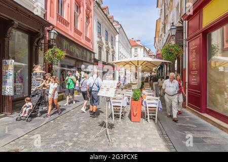 Ljubljana, Slowenien - 15. August 2018: Touristen, die an einem sonnigen Tag in der Altstadt durch die malerische Stari-Straße spazieren, entlang von Restauranttischen Stockfoto