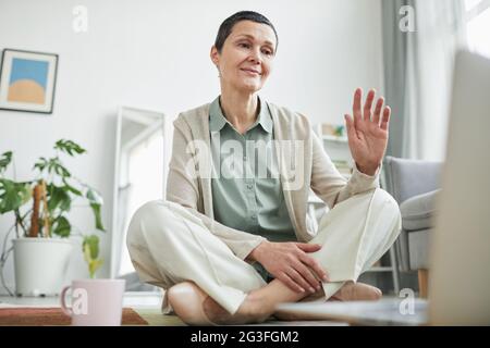 Blick aus der Ferne auf eine lächelnde, reife Frau, die während eines Videoanrufs über einen Laptop in einer gemütlichen Wohnumgebung mit Platz zum Kopieren grüßt Stockfoto