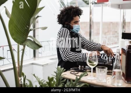 Afrikanischer dj, der in der Cocktailbar im Freien Musik spielt, während er eine Gesichtsschutzmaske trägt - Fokus auf das Gesicht Stockfoto