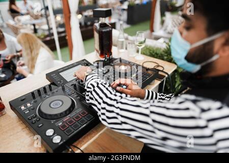 Afrikanischer dj, der in der Cocktailbar im Freien Musik spielt, während er eine Gesichtsschutzmaske trägt - Fokus auf die rechte Hand Stockfoto