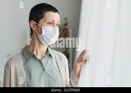 Porträt einer traurigen, reifen Frau, die zu Hause eine Maske trägt und auf ein Fenster schaut, ein kovidges, soziales Distanzierungskonzept Stockfoto