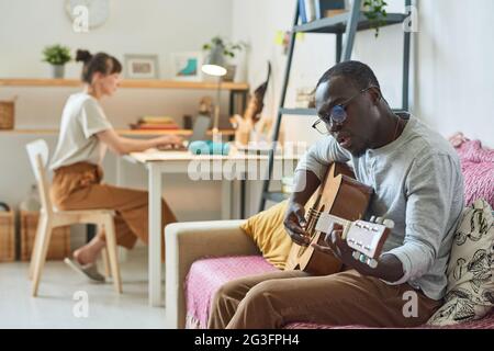 Afrikanischer Mann sitzt auf dem Sofa und spielt Gitarre mit Frau, die im Hintergrund am Tisch sitzt Stockfoto