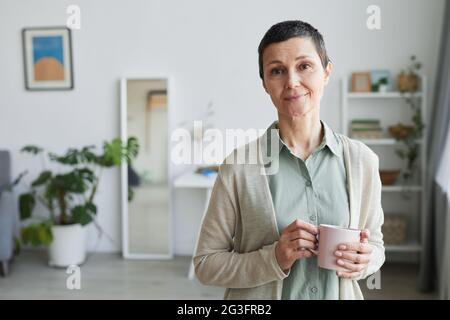 Porträt einer reifen Frau, die am Fenster lächelt, während sie zu Hause eine Tasse Kaffee hält und Platz kopiert Stockfoto