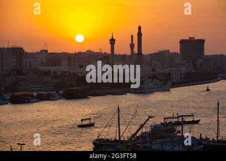 Eine Skyline von Dubai bei Sonnenuntergang mit drei Minaretten in Silhouette und einem großen Gebäude vor dem orangen Himmel. Die Sonne geht unter, Dubai Creek. Stockfoto