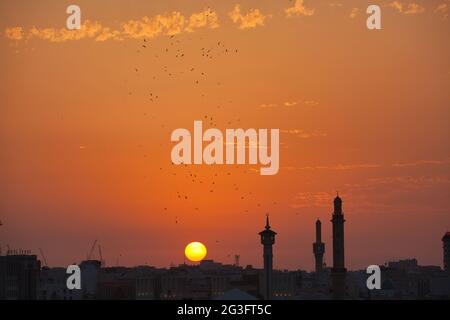 Eine Skyline-Aufnahme von Dubai City bei Sonnenuntergang mit drei Minaretten in Silhouette und einer großen Vogelschar vor dem orangen Himmel. Die Sonne geht unter Stockfoto