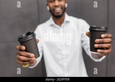 Hübscher und stilvoller afro-amerikanischer lächelnder Mann, der isoliert im Freien steht und zwei Tassen Kaffee zum Mitnehmen vor der Kamera zeigt. Glücklicher Mann, der lächelt Stockfoto