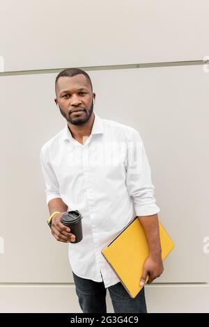 Stilvoller, selbstbewusster afroamerikanischer Mitarbeiter oder Geschäftsmann, der im Freien in legerer Kleidung steht, eine Tasse Kaffee und einen Laptop mit zum Mitnehmen hält und Arbeit hat Stockfoto