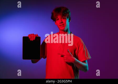 Halten Tablet, verrückt aufgeregt. Porträt eines kaukasischen jungen Mannes auf schwarzem Studiohintergrund in Neonlicht. Schönes männliches Modell in weißem Hemd. Stockfoto
