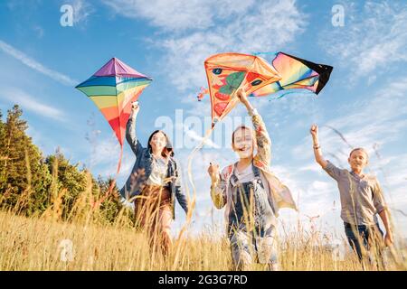 Lächelnde gils und Bruder Junge laufen mit fliegenden bunten Drachen auf der hohen Graswiese in den Bergfeldern. Glückliche Kindheitsmomente oder Outdoor-Ti Stockfoto