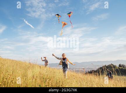 Lächelnde gils und Bruder Junge mit fliegenden bunten Drachen - beliebtes Outdoor-Spielzeug auf der hohen Graswiese in den Bergfeldern. Glückliche Kindheitsmomente Stockfoto