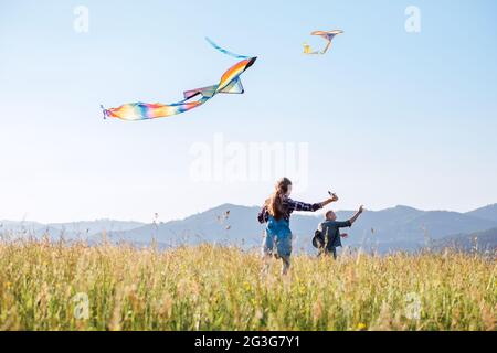 Die kleine Tochter mit dem Vater läuft schnell, während sie bunte Drachen auf der hohen Graswiese in den Bergfeldern auffliegen. Warme Familienmomente Stockfoto