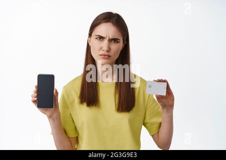 Porträt einer jungen Frau zeigt Kreditkarte und Mobiltelefon, verwirrt runzelig, verwirrt über Zahlungssystem, seltsame Sache auf dem Bankkonto, stehend Stockfoto