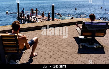 Kiel, Deutschland. Juni 2021. Zwei junge Männer blicken von ihren Liegestühlen auf Badegäste, die von einer Badeplattform in einen zum Schwimmen gesperrten Bereich der Kieler Förde springen. Auch im Norden beginnt der Sommer mit großer Hitze. Quelle: Axel Heimken/dpa/Alamy Live News Stockfoto