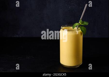 Mango Lassi aus Glas mit Rohr auf dunklem Hintergrund. Traditionelles indisches und pakistanisches Getränk mit Joghurt, Mango, Gewürzen, Minze und Eis Stockfoto