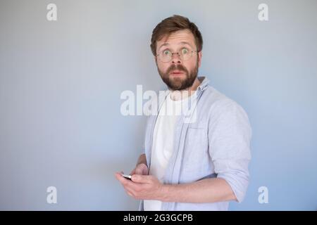 Verwirrt, schockiert Mann mit einem Smartphone in den Händen schaut auf die Kamera überrascht, mit großen Augen. Grauer Hintergrund. Stockfoto