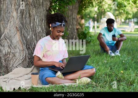 Afrikanische junge Frau, die im Park auf dem Rasen sitzt und online am Laptop arbeitet Stockfoto
