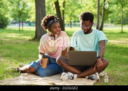 Afrikanisches junges Paar, das auf dem Gras sitzt, Kaffee trinkt und im Park einen Laptop benutzt Stockfoto
