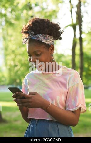 Afrikanische glückliche Frau, die online mit ihrem Mobiltelefon spricht, während sie im Park steht Stockfoto