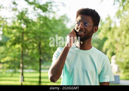 Afrikanischer junger Mann in einer Brille, der im Park steht und am Handy spricht und lächelt Stockfoto