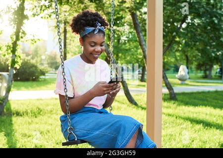 Glückliche Afrikanerin, die auf einer Schaukel sitzt und im Park eine Nachricht auf dem Handy tippt Stockfoto