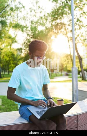 Afrikanischer junger Mann, der auf dem Laptop tippt und online arbeitet, während er im Park sitzt Stockfoto