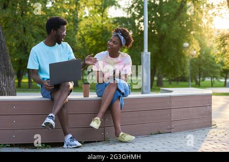 Afrikanisches junges Paar, das auf der Bank im Park mit einem Laptop sitzt und spricht Stockfoto