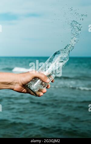 Nahaufnahme eines Mannes, der Wasser aus einer wiederverwendbaren Glasflasche vor dem Ozean ausgießt Stockfoto
