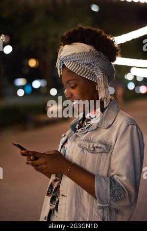Afrikanische junge Frau, die am Abend online über das Mobiltelefon spricht, während sie in der Stadt spazierengeht Stockfoto