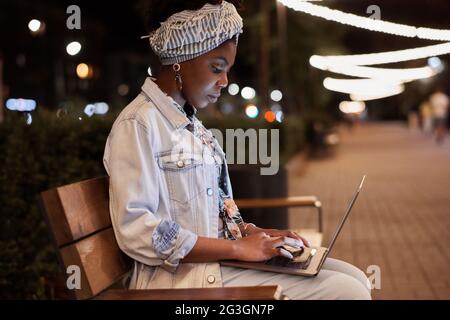 Afrikanische junge Frau, die abends auf der Bank sitzt und auf dem Laptop im Park tippt Stockfoto