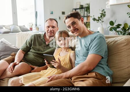 Porträt einer glücklichen, schwulen Familie, die lächelt, während sie mit dem Kind auf dem Sofa sitzt und einen Tablet-pc benutzt Stockfoto