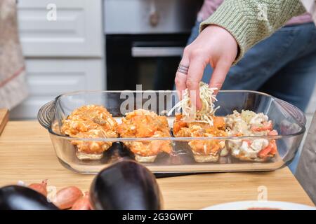 Nicht erkennbare junge Frau, die Käse zu den gefüllten Auberginen hinzufügt. Garzeit. Stockfoto