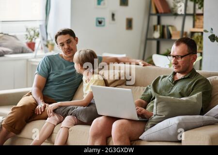 Vater spricht mit Tochter, während sie auf dem Sofa im Zimmer sitzen und ein anderer Mann am Laptop arbeitet Stockfoto