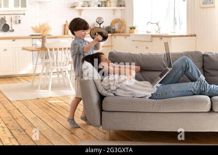 Gestresste Mutter decken Ohr von lauten Sohn bang Geschirr. Müde von einer jungen Frau, die mit geschlossenen Augen auf dem Sofa lag Stockfoto