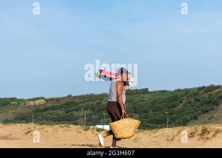 Ein Algerier mittleren Alters, der einen traditionellen französischen Marktkorb und Teppiche am Strand hält. Stockfoto