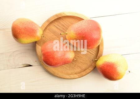 Mehrere reife gelb-rote Birnen mit rundem Holztablett, Nahaufnahme, auf einem weißen Holztisch, Draufsicht. Stockfoto