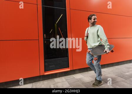 Fröhlicher Mann im Sweatshirt, der Longboard hält und an der orangen Wand entlang geht Stockfoto