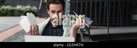 Bärtiger Mann in Polohemd und Sweatshirt, der Essstäbchen und Karton hielt, während er im Café im Freien einen Laptop ansah, ein Banner Stockfoto