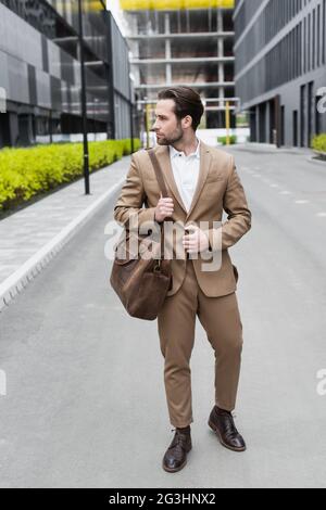 In voller Länge bärtiger Geschäftsmann in Anzug, der Lederriemen der Tasche draußen hält Stockfoto