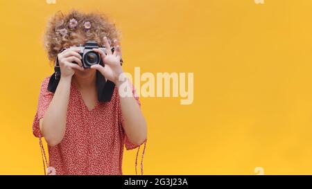 Schönes Mädchen mit lockigen Haaren ein Bild machen. Isoliert über gelbem Hintergrund Studio. Attraktives Mädchen in rosa Top gekleidet, trägt Blumen in den Haaren und hält eine Kamera. Fotokonzept. Stockfoto