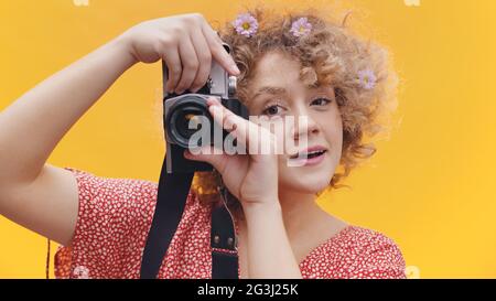 Fröhliches Mädchen, das eine Kamera in den Händen hält und ein Foto fotografiert. Isoliert über gelbem Hintergrund Studio. Attraktive Mädchen in rosa Top gekleidet trägt Blumen in den Haaren. Fotokonzept. Stockfoto