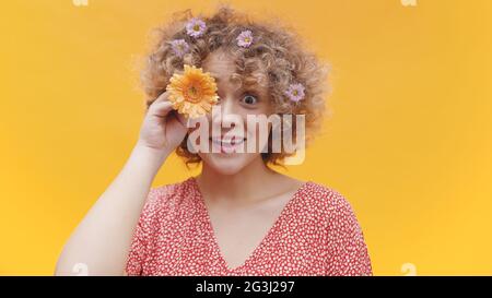 Schönes junges Mädchen, das ihr Auge mit einer orangefarbenen Gerbera-Gänseblümchen bedeckt. Isoliertes Mädchen mit hellgelben Hintergrund Studio. Funky Mädchen lächelt und trägt Blumen in ihrem lockigen Haar. Stockfoto