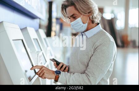 Männlicher Reisender mit Gesichtsmaske, der den Check-in mit einem Automaten am Flughafenterminal macht. Reifer Mann, der am Flughafen selbst einchecken kann. Stockfoto