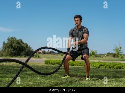 Männlicher Athlet im Freien im Park, Übungen für Ausdauer, mit einem dicken Seil, für Cross-Fitness. Europäisch-Kaukasisch Stockfoto