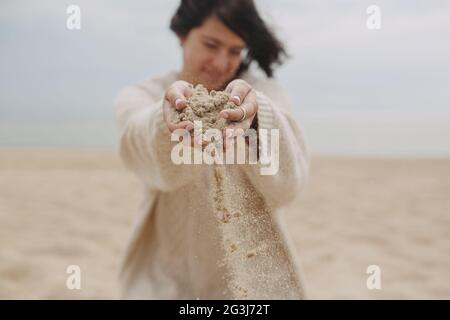 Stilvolle junge Frau im Pullover Freigabe Sand am Strand, die Hände aus nächster Nähe. Sand läuft durch die Hände. Zeitkonzept. Sommer Strand und Urlaub. Frau han Stockfoto